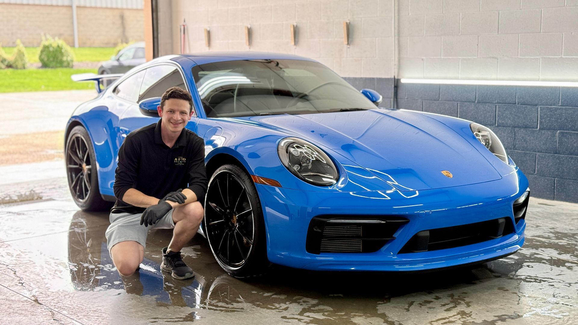 Man crouches beside a bright blue sports car being washed in a driveway.