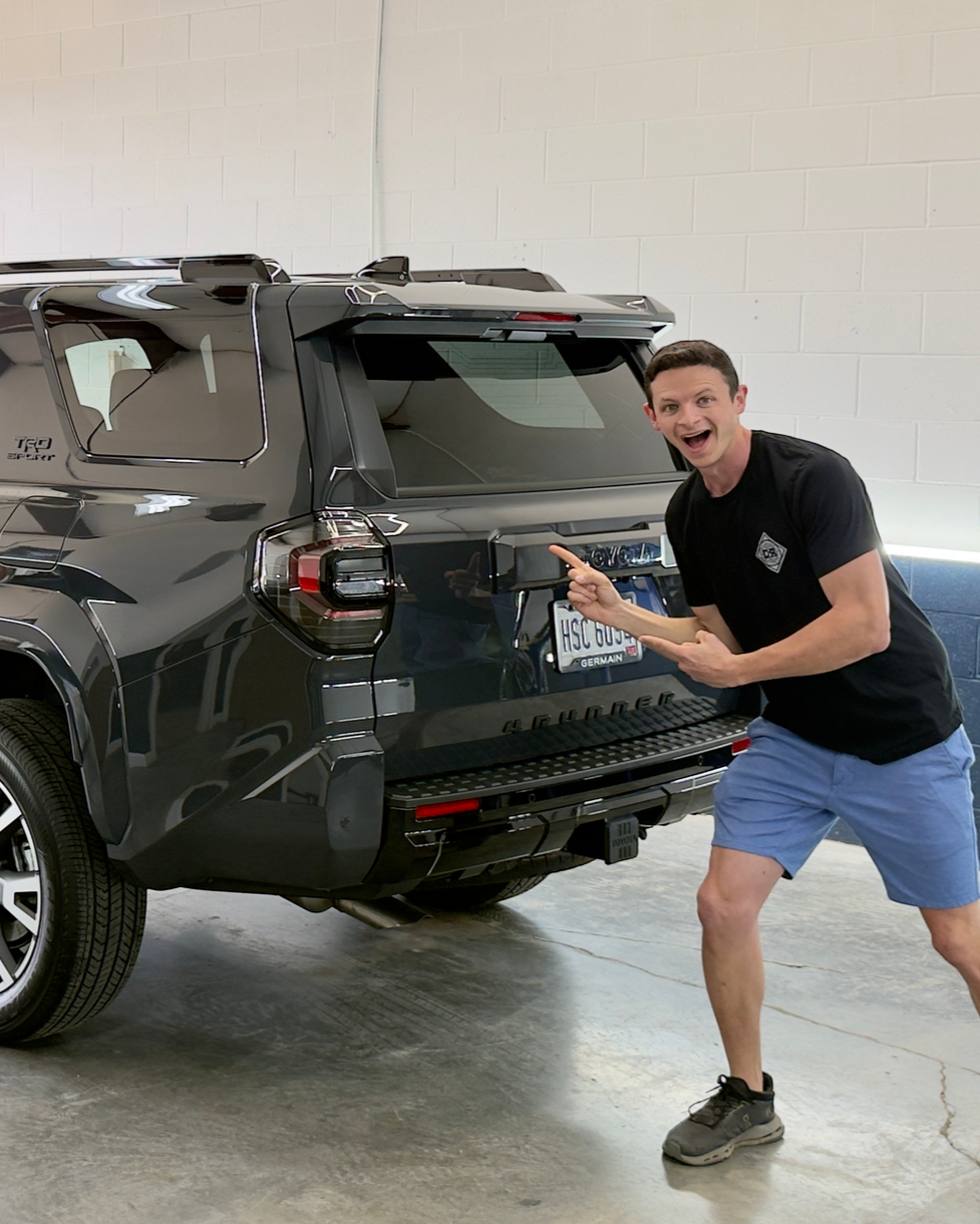 Two men smile for a selfie in an auto garage, one waving and the other making a hand gesture, with cars in the background.
