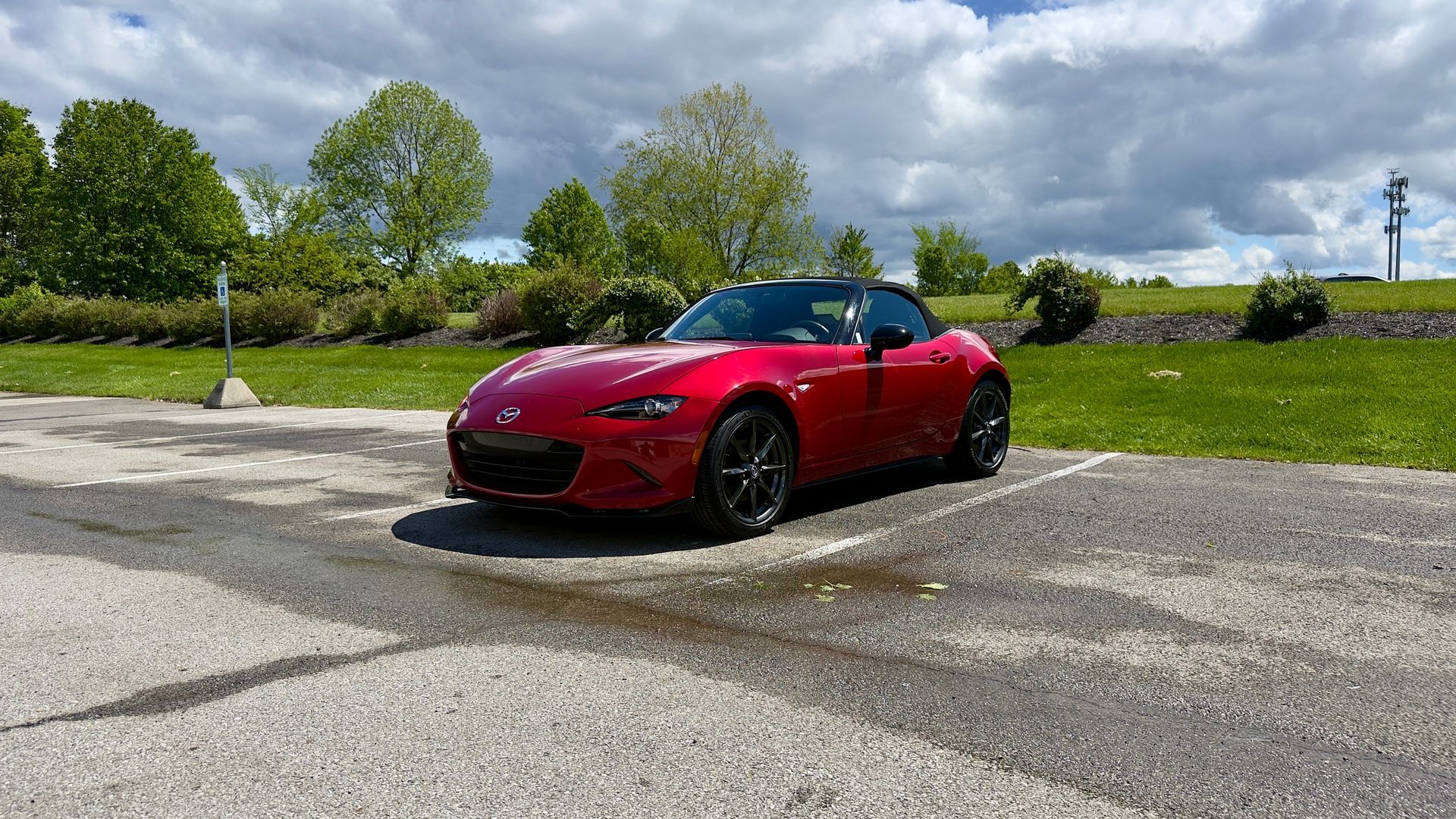 Red convertible sports car parked on a wet lot under a cloudy sky, with trees and grass in the background