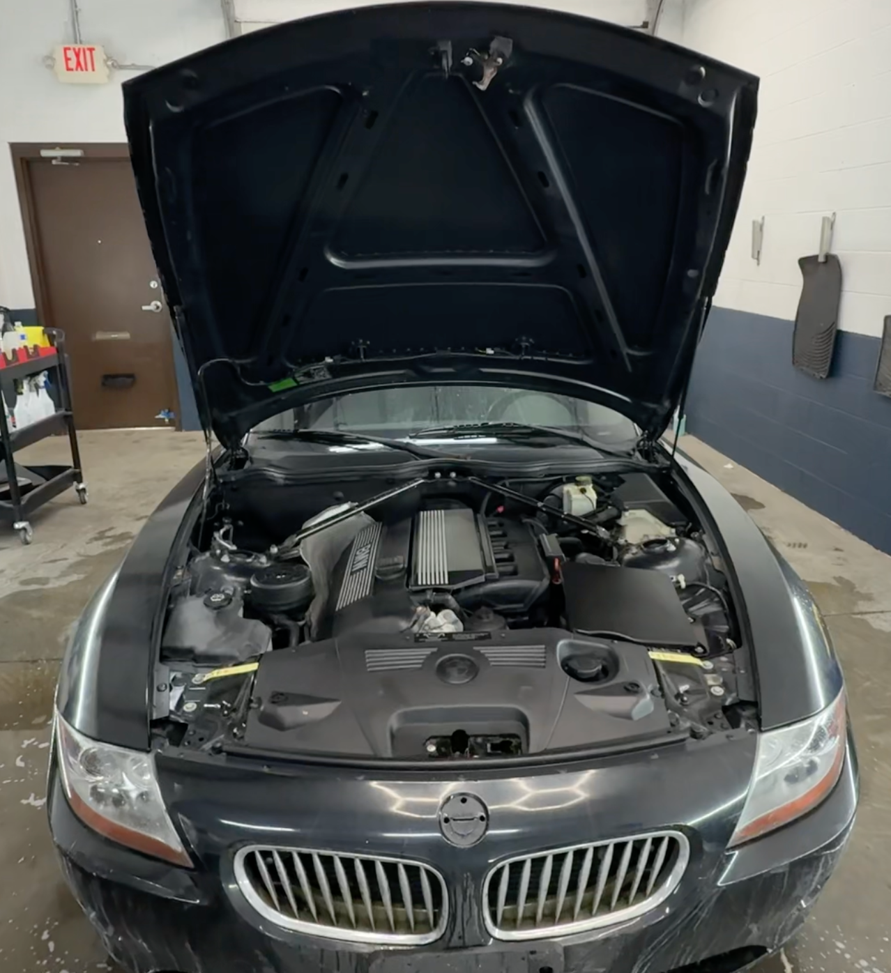 Black BMW sedan with hood open inside an auto shop, engine bay exposed.