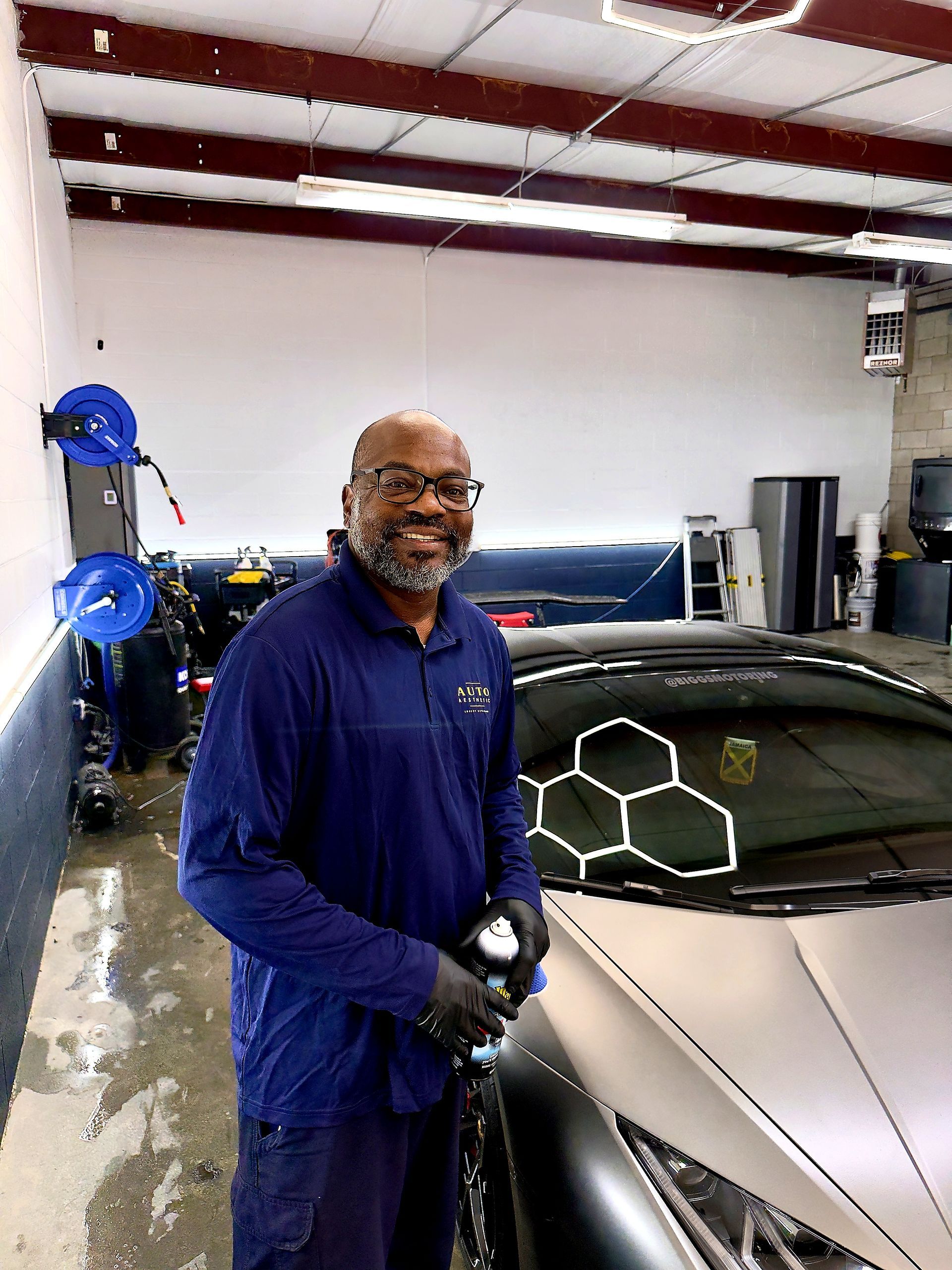 Man in blue coveralls smiling beside a silver sports car in a garage