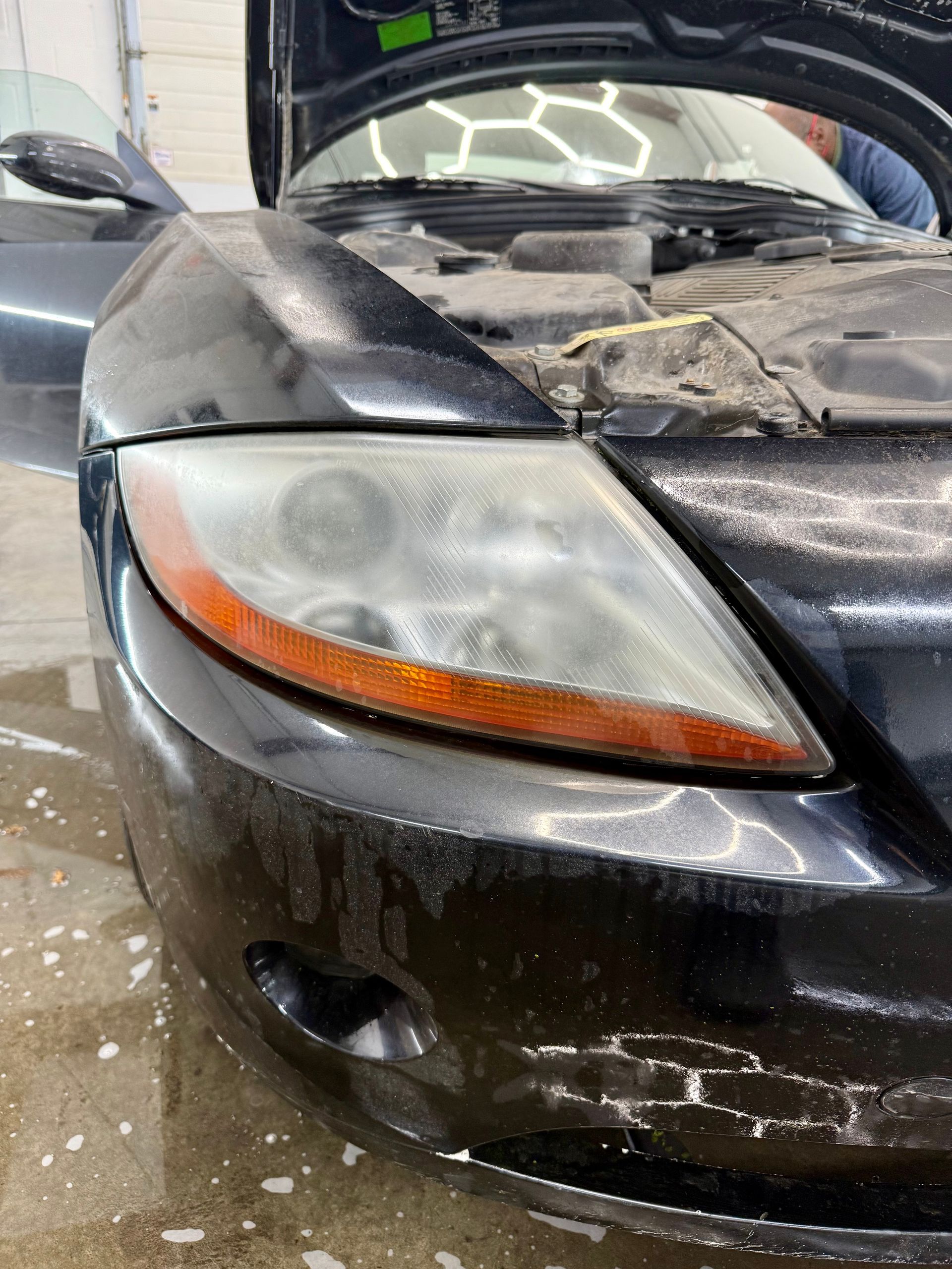 A close-up view of the front headlight and bumper of a black car with some water spray on a concrete surface.
