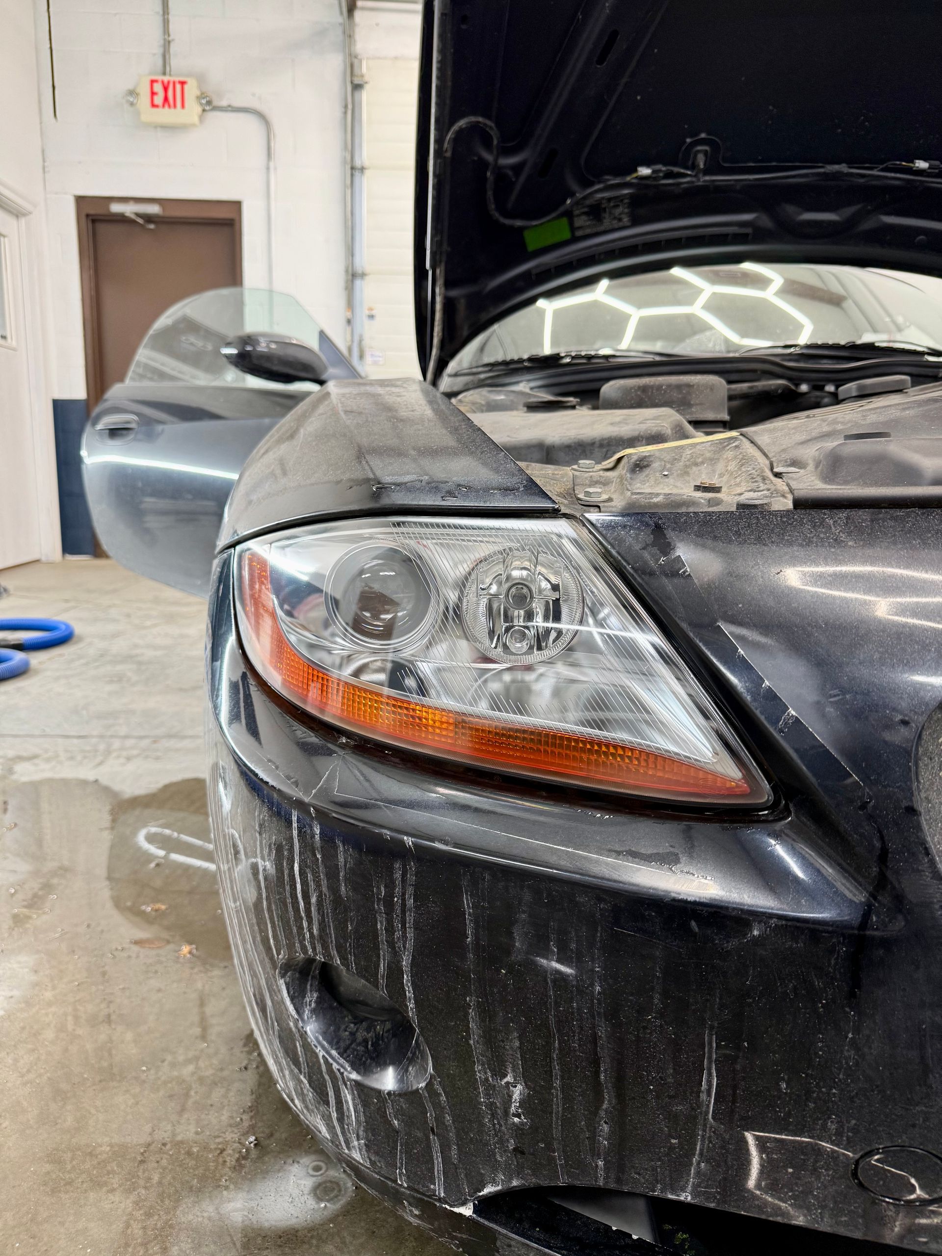 Close-up of a black car's front passenger headlight and bumper, covered in white cleaning foam inside a garage.