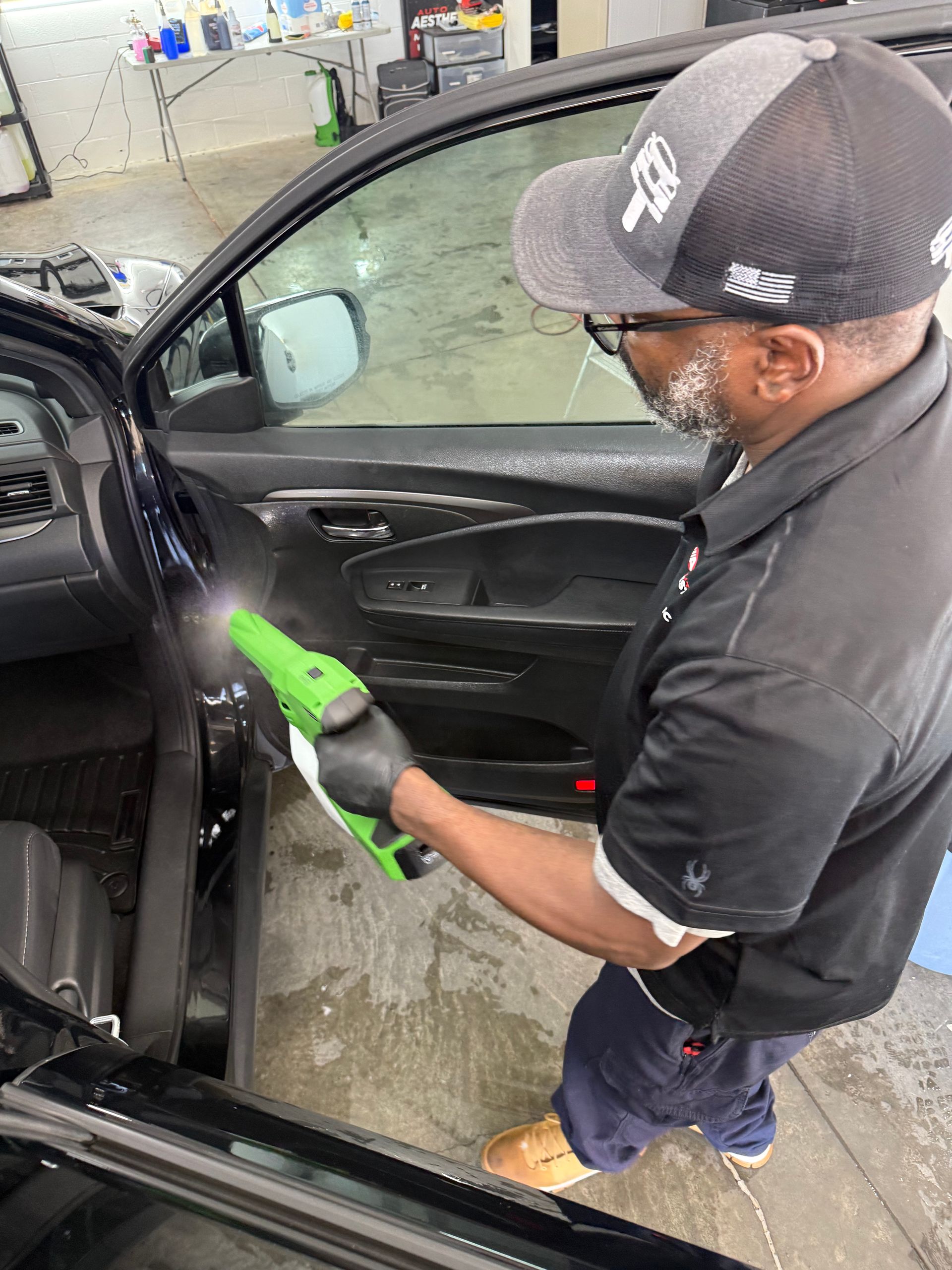 A person wearing a grey cap and black polo cleans the interior door panel of a car using a green handheld spray tool.