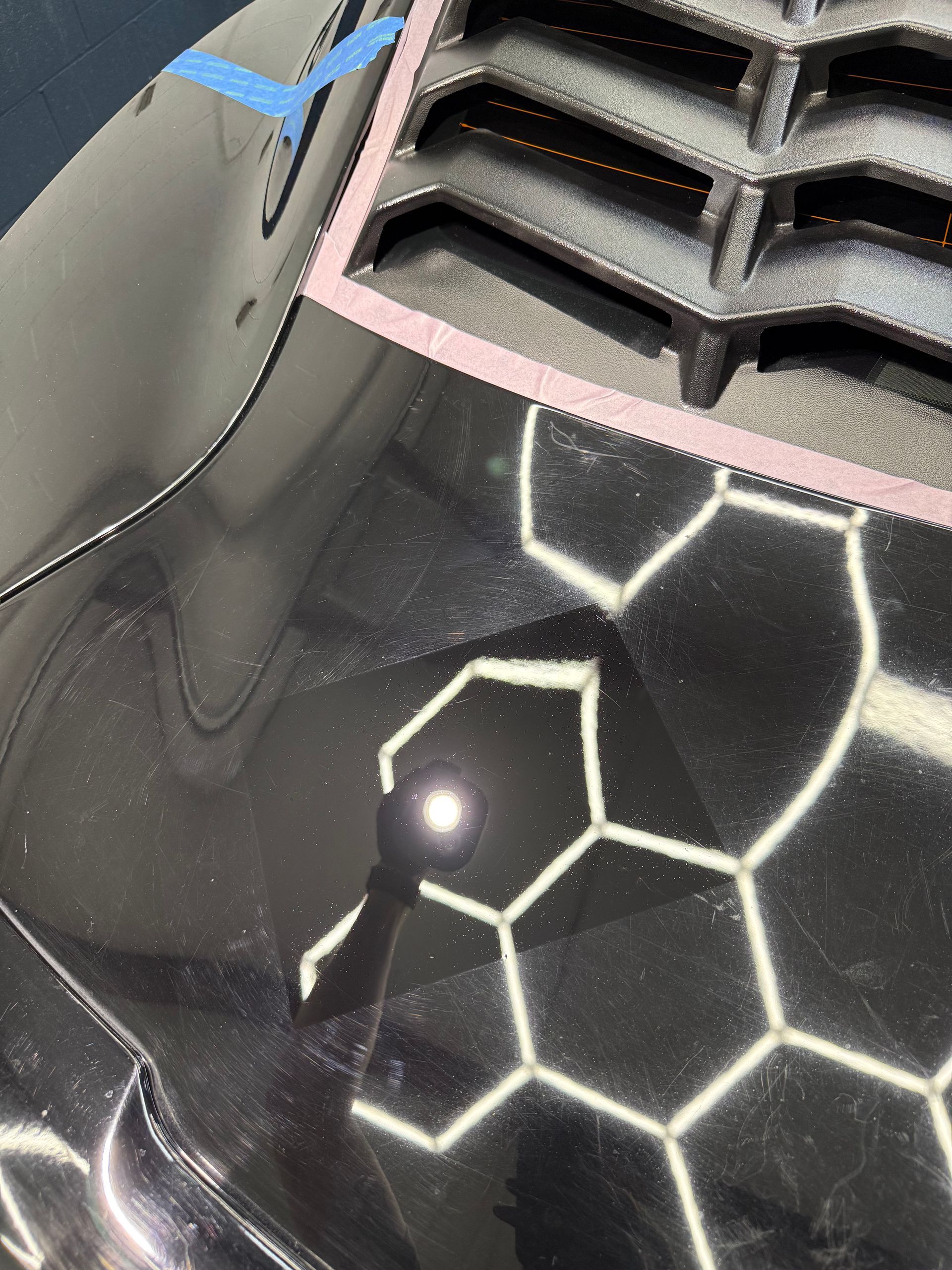 Close-up of a black car hood with white hexagon pattern and blue tape near the headlight grille