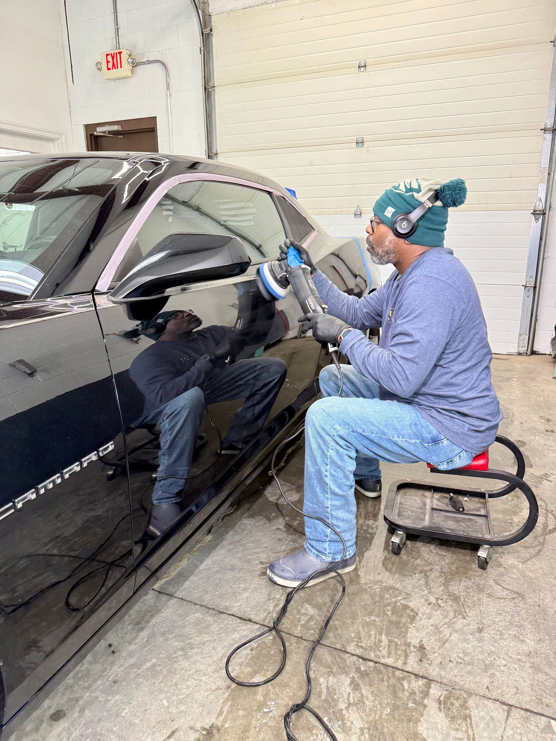 Person polishing a black car in a garage, using a polisher while seated on a stool.