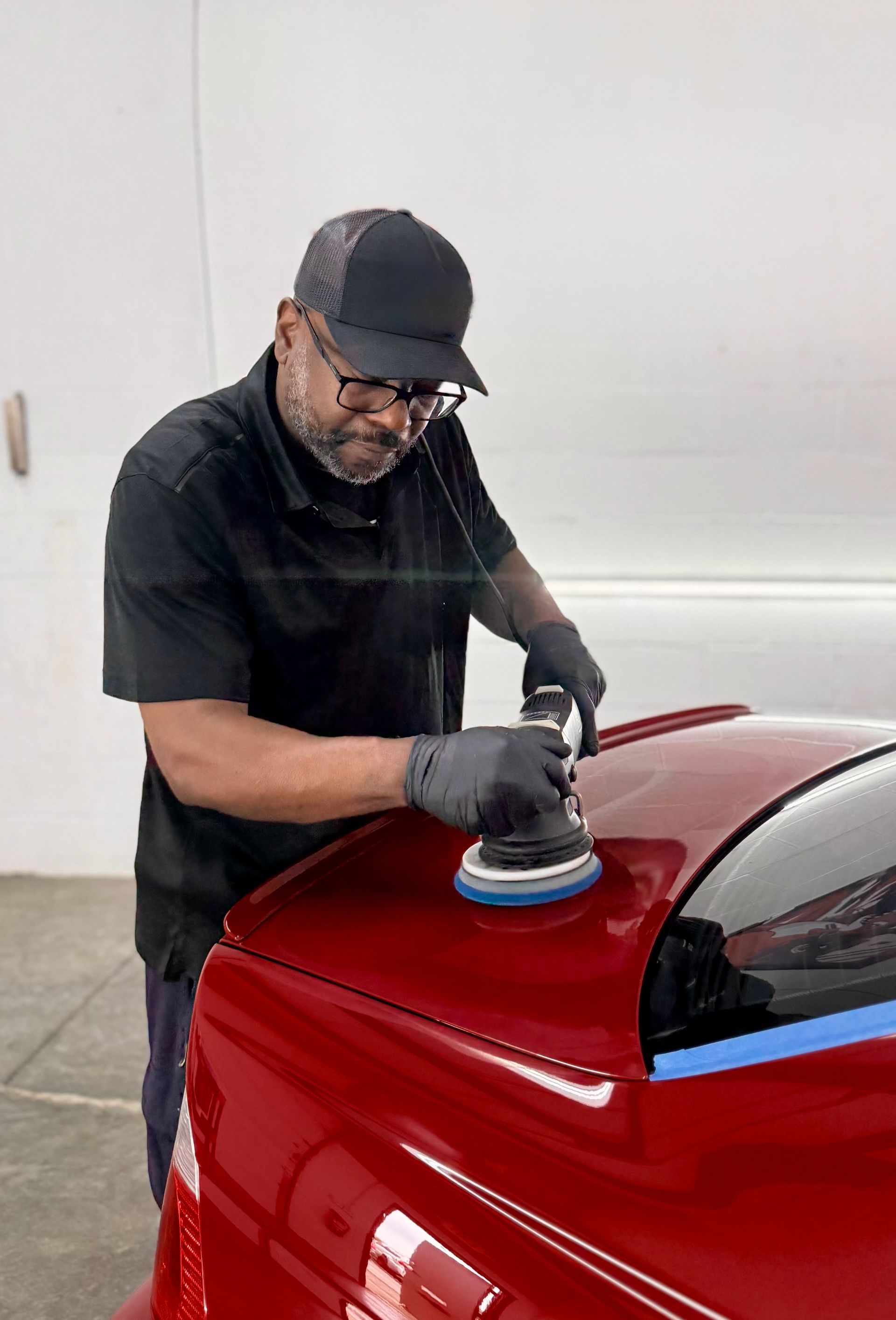 Man polishing a red car in a well-lit shop, wearing a black cap, shirt, gloves, and glasses.