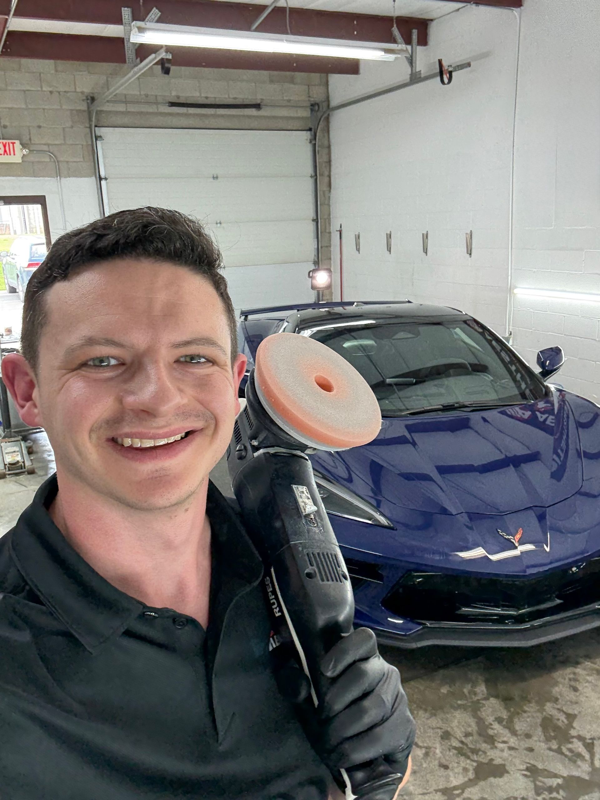 Man holding buffer in front of blue sports car, smiling, in a car detailing shop.