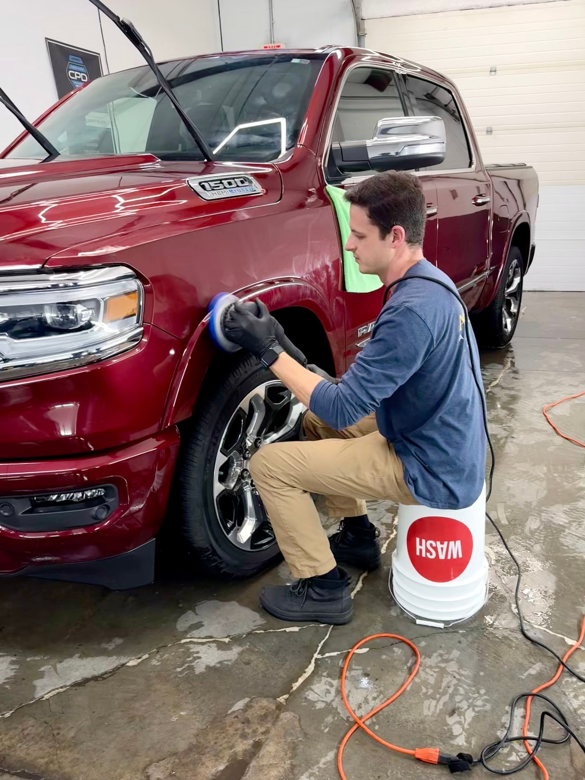 Man using a polisher on a red truck. He sits on a bucket in a garage.