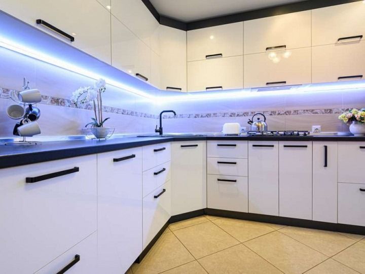 Modern white kitchen with black countertops and cabinet handles, blue LED lighting, and beige tile floor.