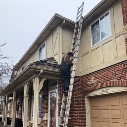 Person on tall ladder climbing side of a two-story brick and beige house. Overcast sky.