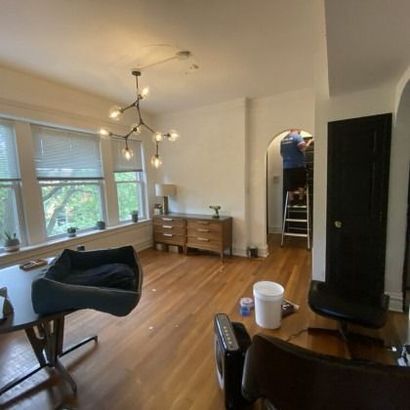 A room with a dog bed, table, chest of drawers, and person on a ladder in an arched doorway.