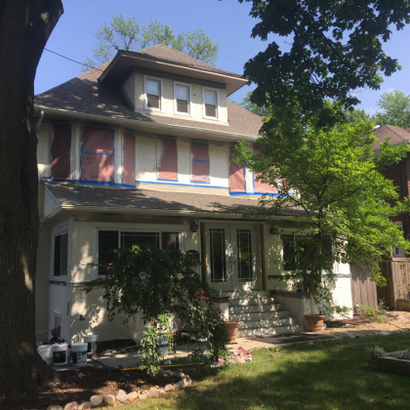 Two-story house undergoing renovation with windows covered. Beige stucco, green trees, blue sky.
