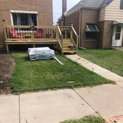 Newly built wooden deck with stairs in front of a brick house. Lumber stacked on the grass below.