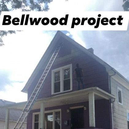 Person painting the purple exterior of a house, standing on a porch. A ladder leans against the roof.