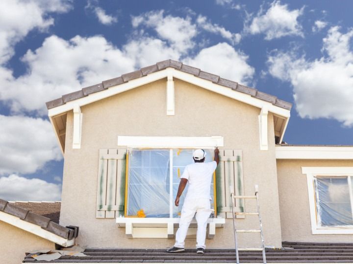 Person in white painting exterior of a house, standing on a ladder. Sky with clouds in the background.