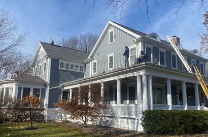 Two workers on a light blue house roof near a ladder. White porch and siding, blue sky.