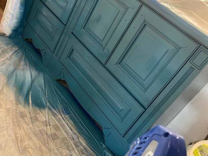 Teal-painted wooden cabinet being sprayed with paint, covered in plastic, indoors.