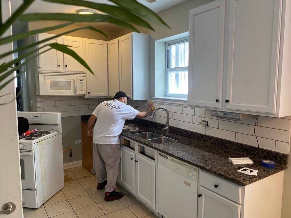 A person cleaning a kitchen countertop with white cabinets. Tile backsplash. Window. Microwave above oven.