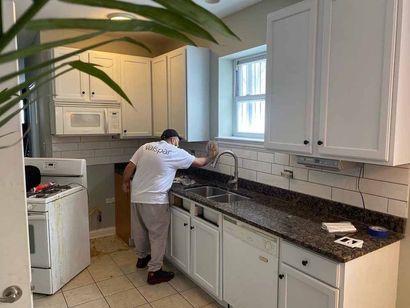 A person cleaning a kitchen countertop with white cabinets. Tile backsplash. Window. Microwave above oven.