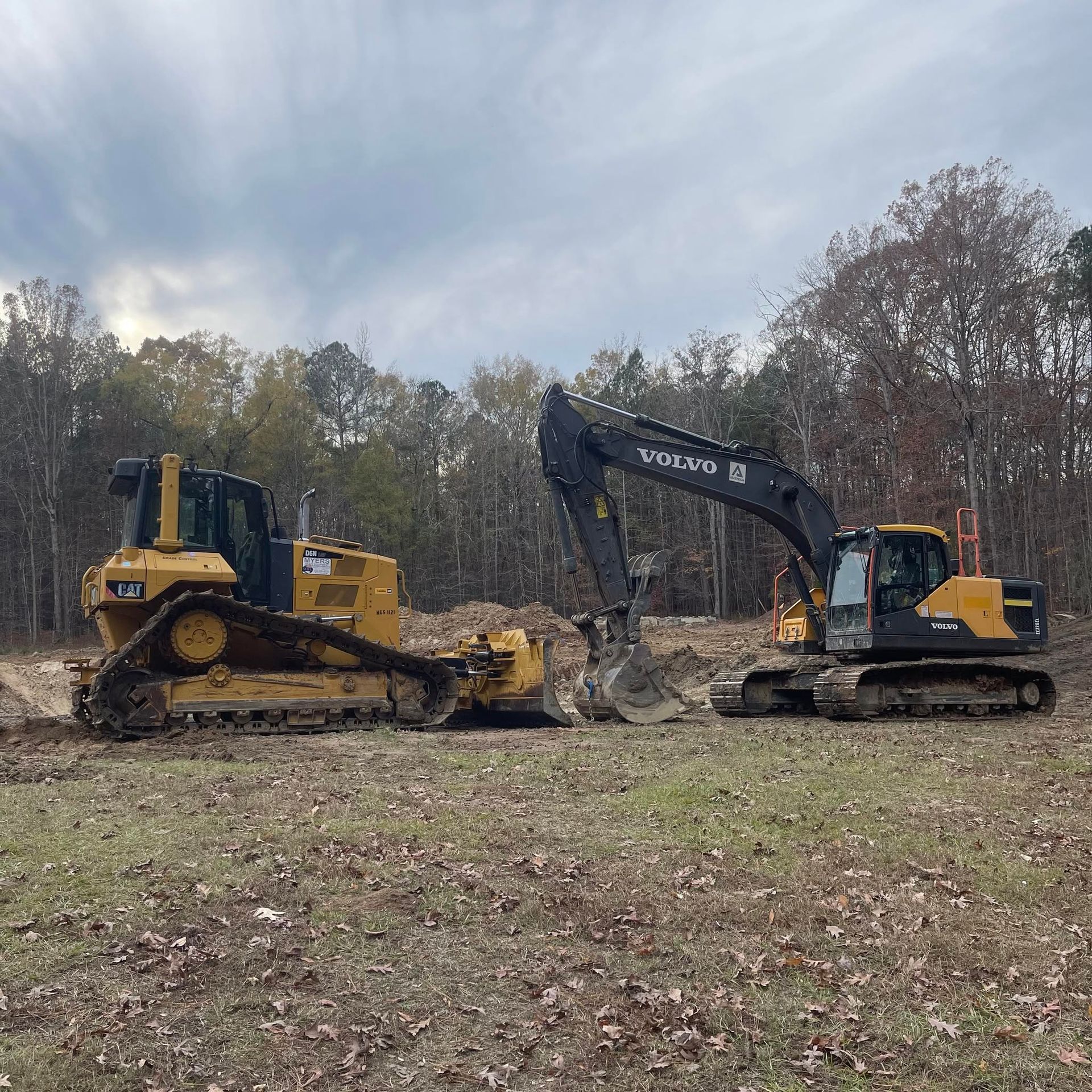 Two tracked excavators and a bulldozer on a grassy field under cloudy sky, with trees in the background