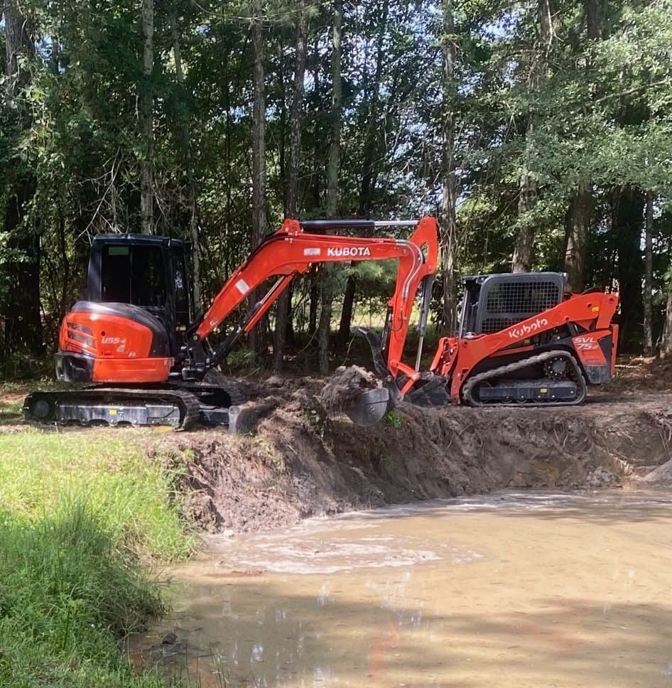 Two orange excavators working beside a muddy pond in a wooded area
