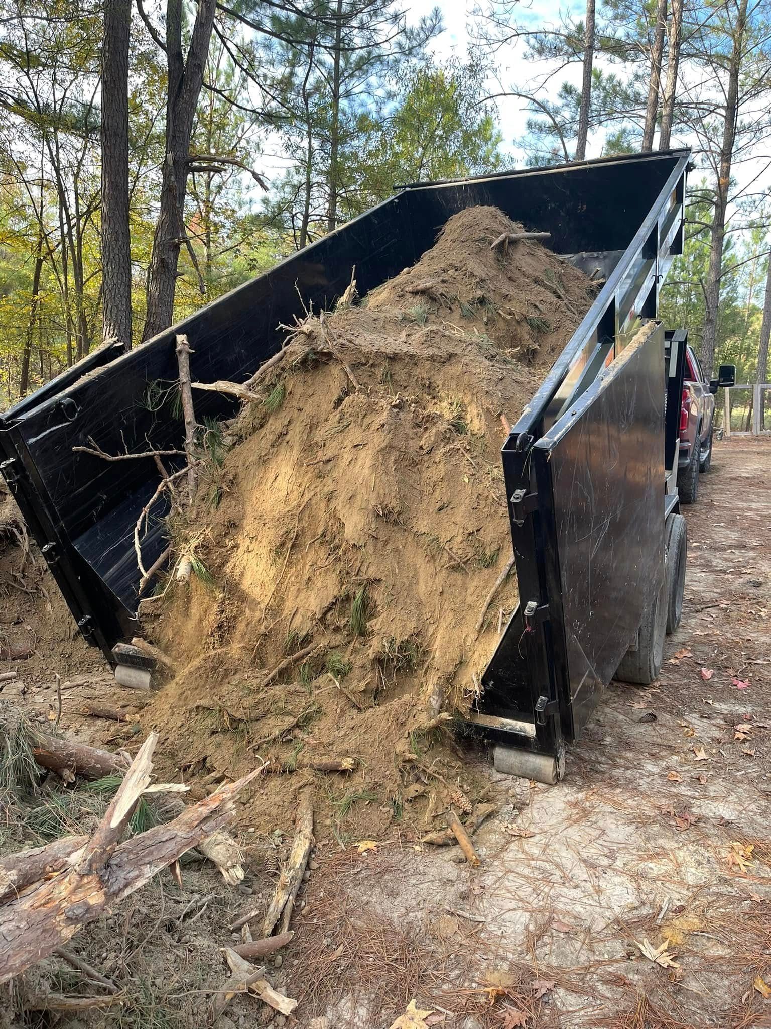 Black dump trailer tipped with a large pile of dirt and roots spilling out in a wooded area