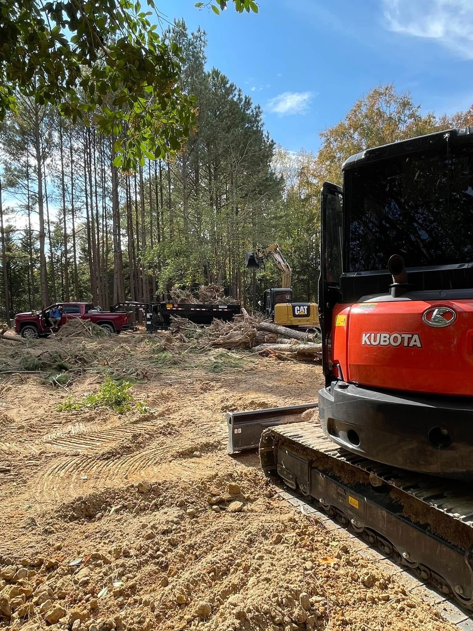 Red Kubota excavator beside a cleared wooded lot with piles of debris under a blue sky