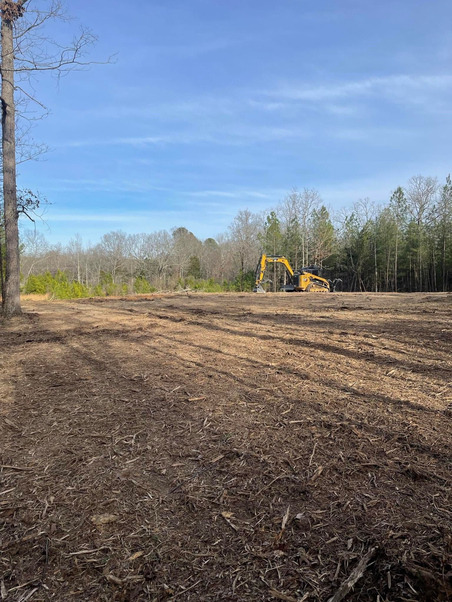 Cleared field with excavator working near tree line under a blue sky