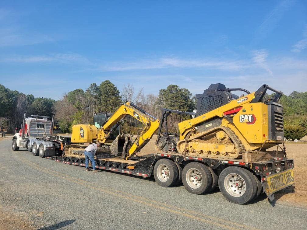Yellow CAT excavators loaded on a flatbed truck on a gravel roadside under a blue sky.