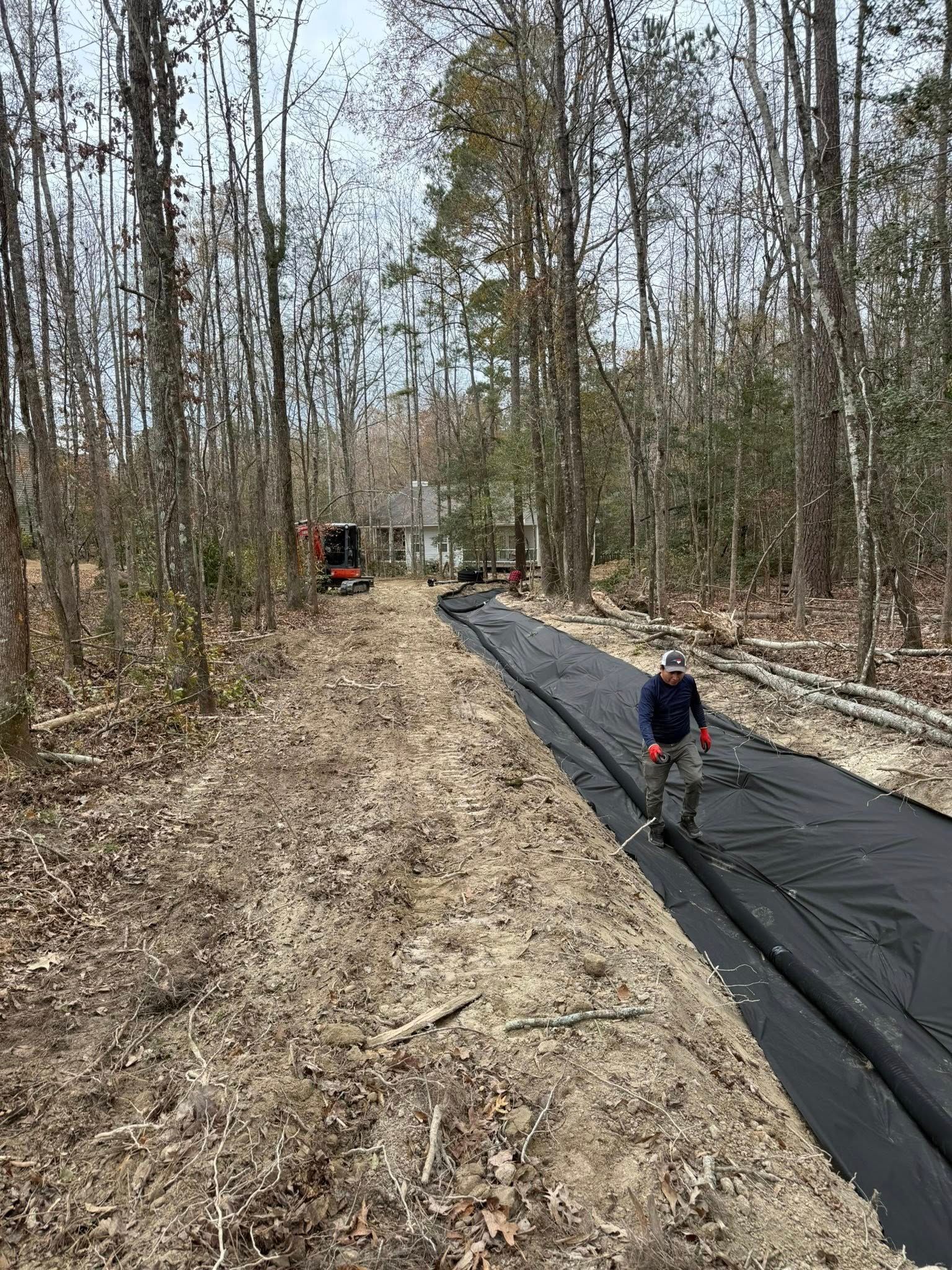Person working on black landscaping fabric along a wooded dirt road with a tractor in the distance