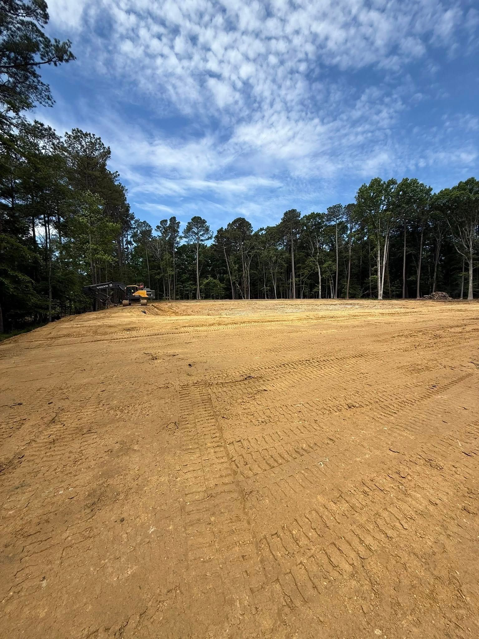 Cleared dirt lot bordered by trees under a blue sky with clouds