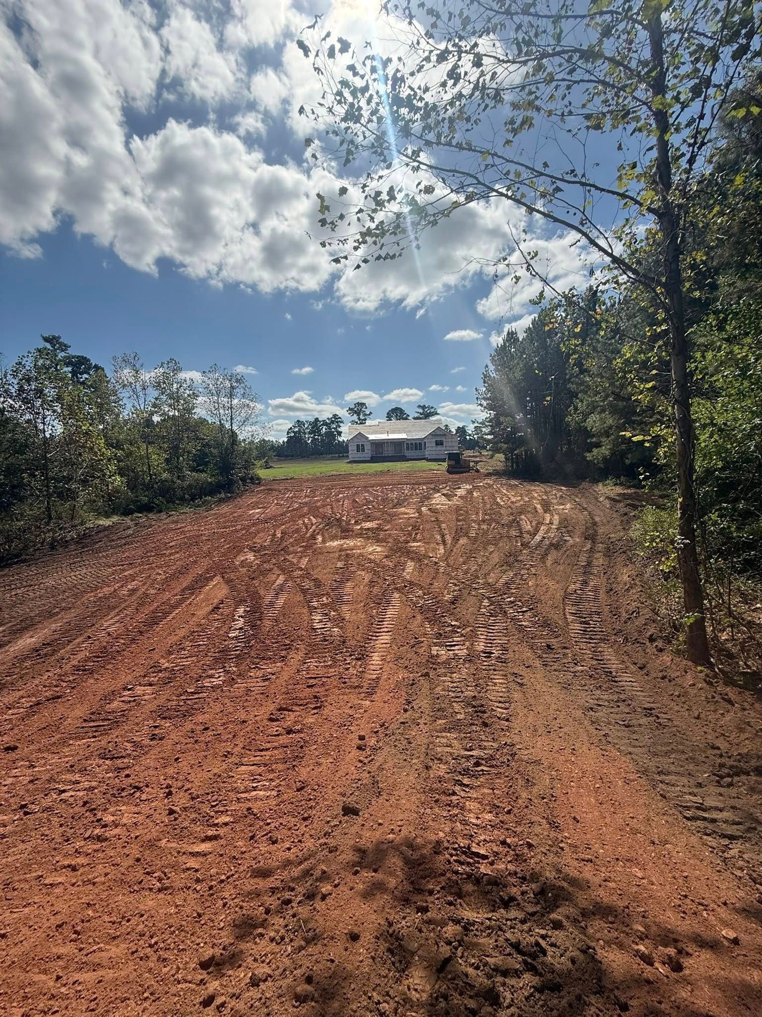 Reddish dirt road under blue sky and clouds, lined with trees and leading to distant houses