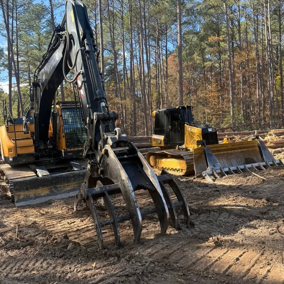 Excavator with grapple and bulldozer blades on a dirt lot in a pine forest