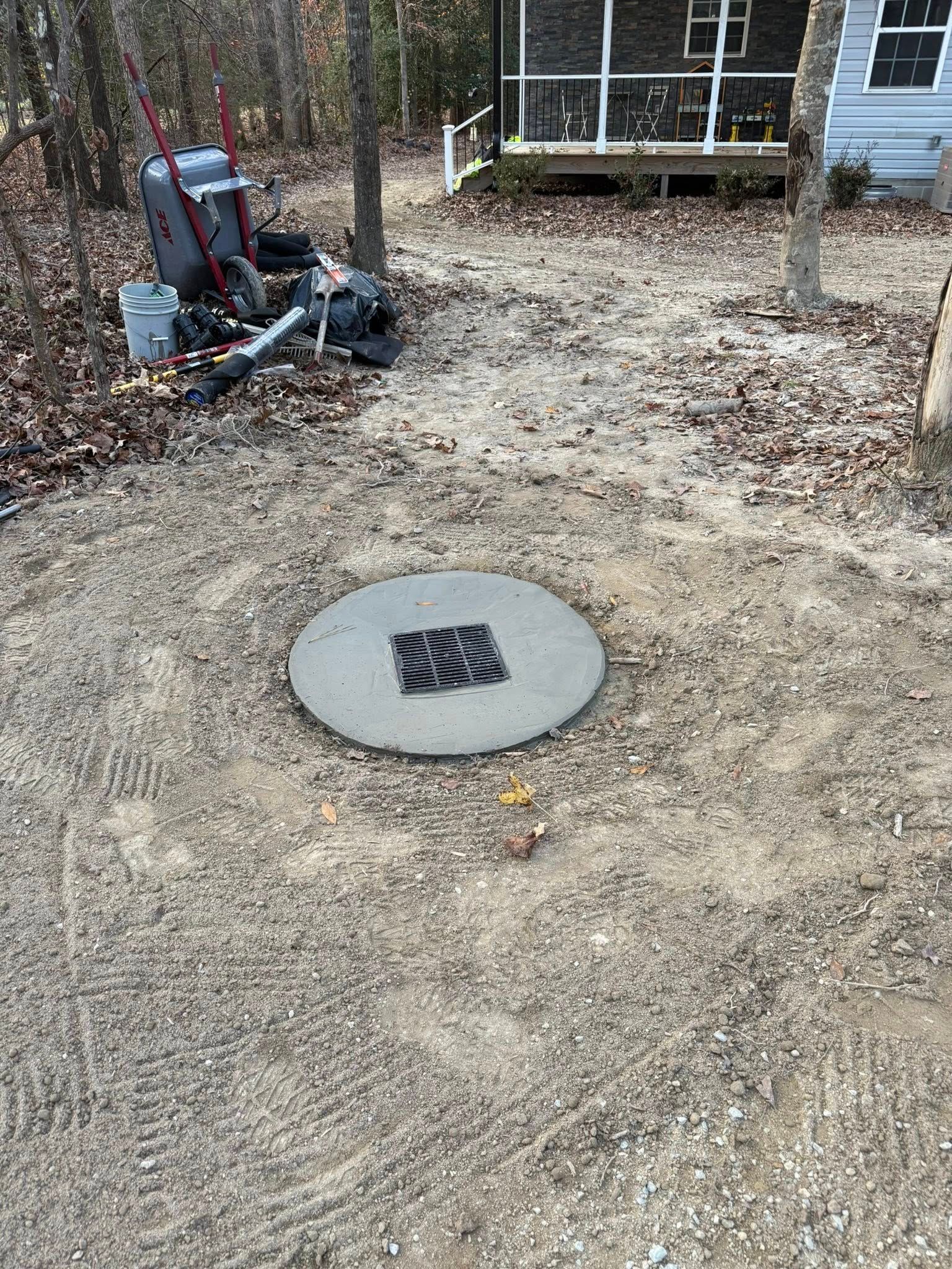 Ground-level view of a round drain cover in a dirt yard, with a wood deck and equipment in the background