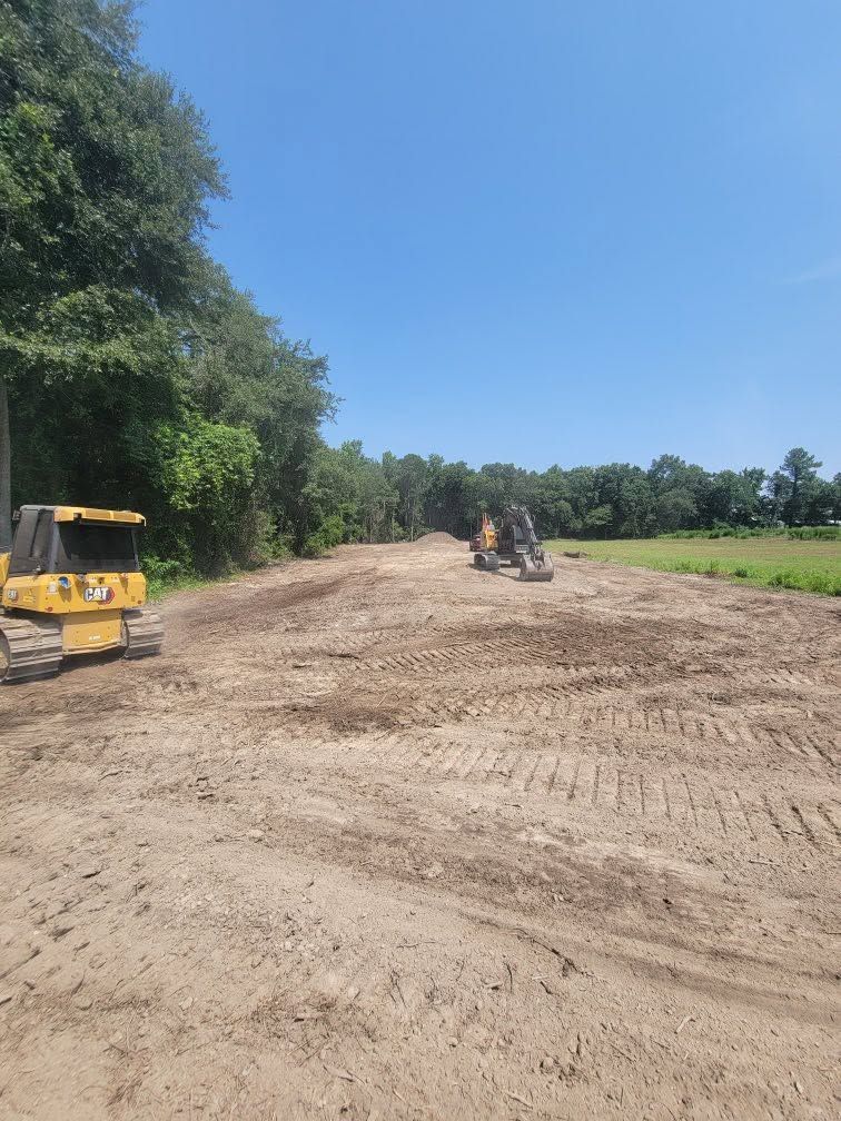 Construction equipment grading a dirt road under a clear blue sky, with trees and open fields nearby