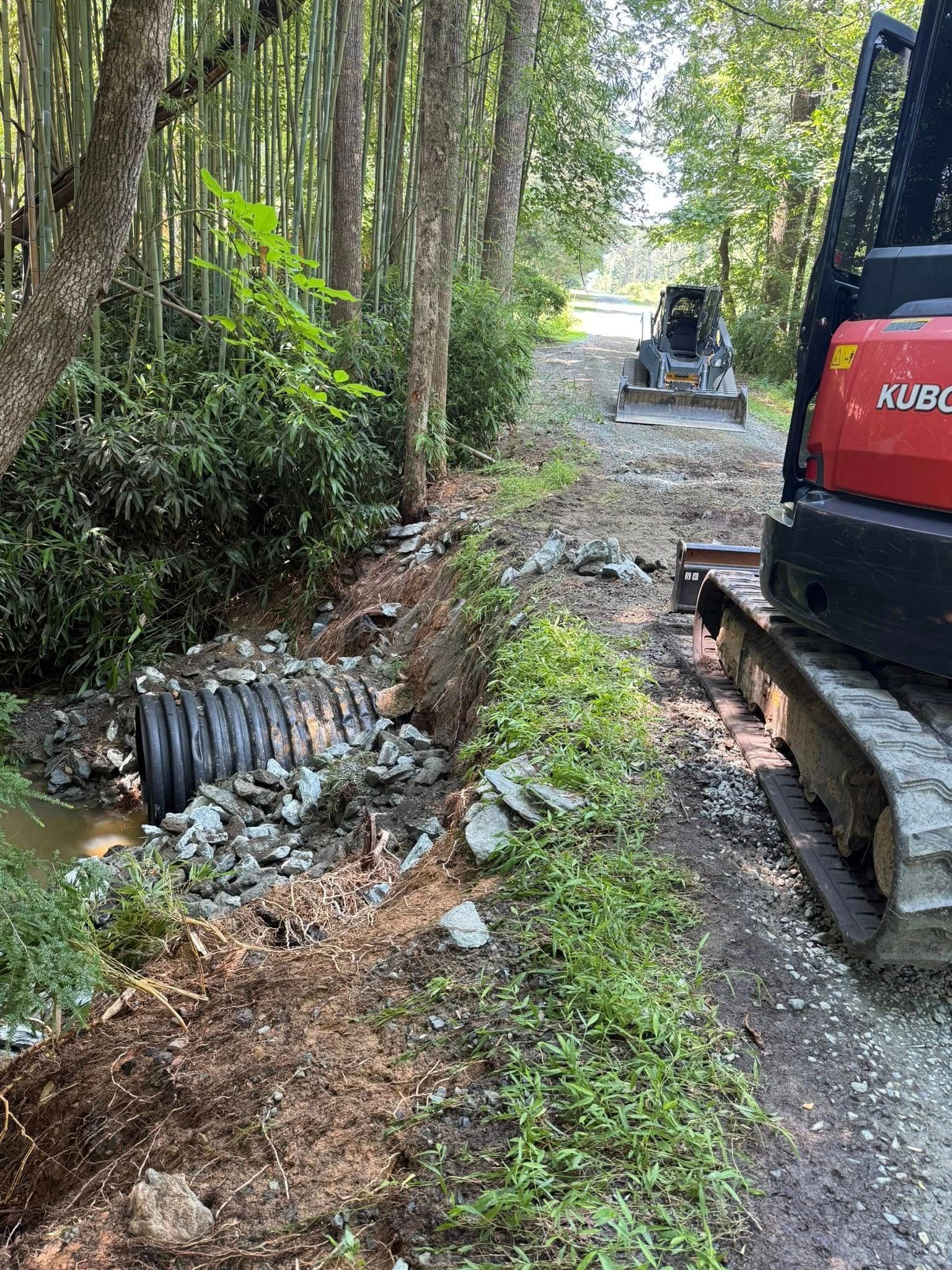 Red excavator on muddy woodland trail beside a damaged culvert and fallen pipe