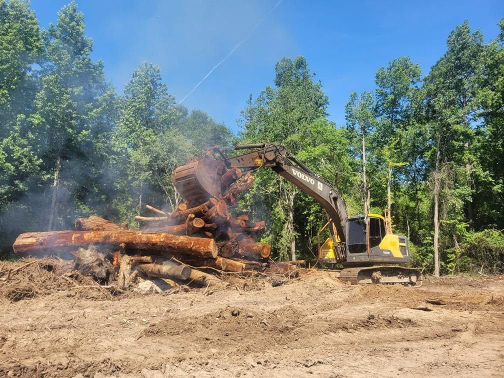Excavator clearing a pile of burning logs in a wooded clearing under a blue sky