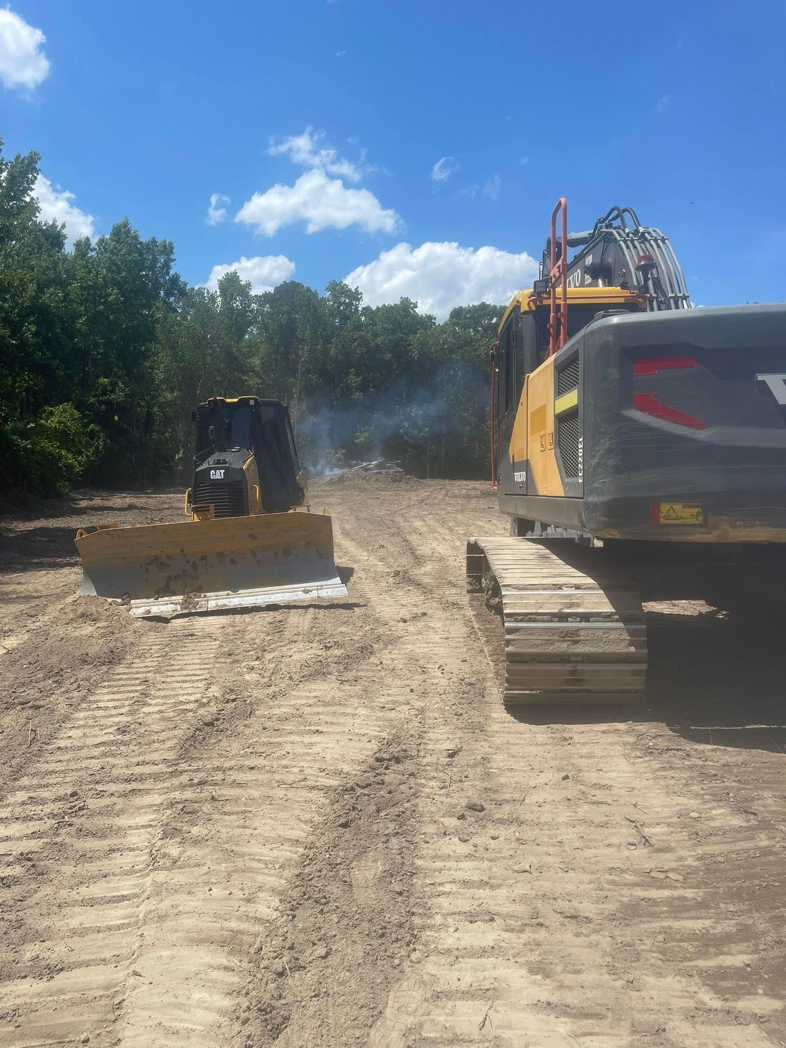 Bulldozer and excavator clearing dirt on a sunny construction site with trees and blue sky