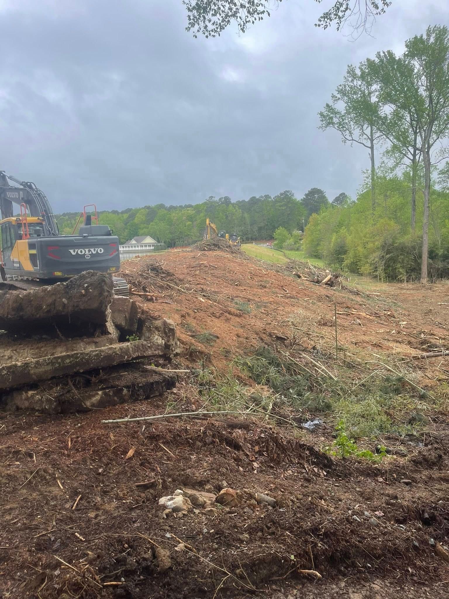 Excavator clearing a muddy hillside under cloudy skies, with trees and a dirt pile nearby