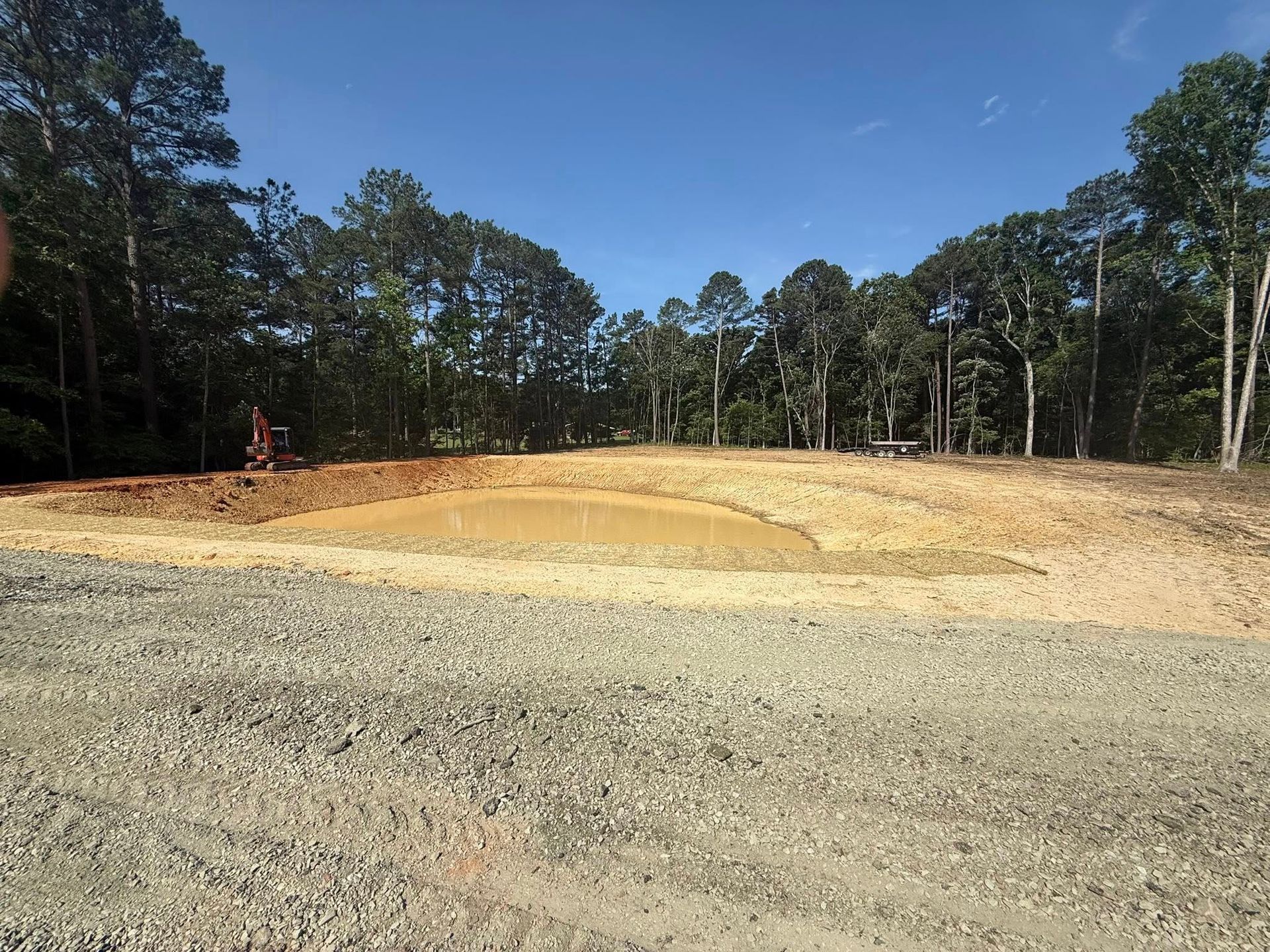 Cleared dirt lot with gravel foreground, trees in the background, and a small excavator on the left