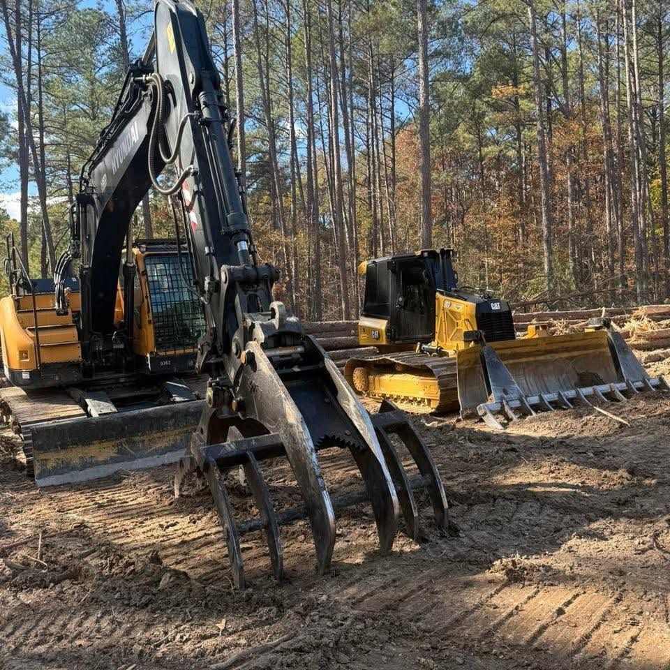 Yellow excavator with grapple attachment on a dirt site beside a bulldozer in a pine forest