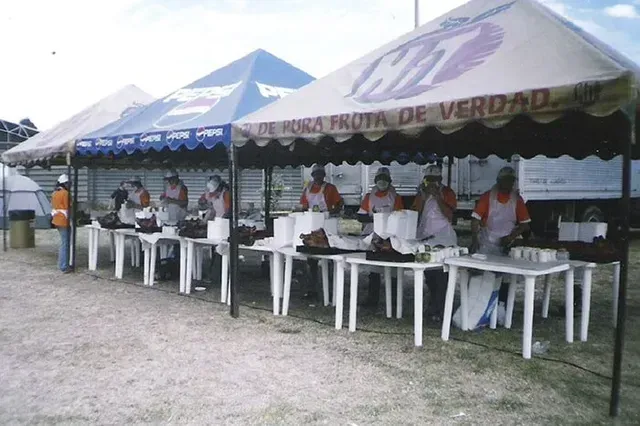 Personas sentadas en mesas largas bajo carpas azules y blancas en un evento al aire libre