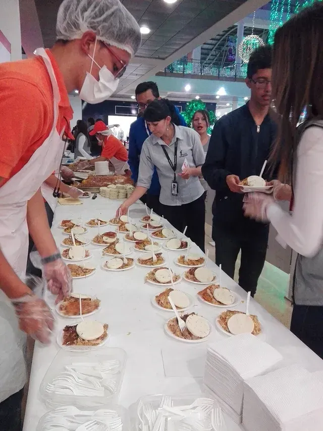 Personas sirviendo muestras de postres a los asistentes en una mesa de bufé, en un salón interior concurrido