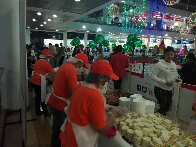 Voluntarios con camisetas naranjas sirviendo comida en un evento multitudinario en un espacio cerrado, con montones de vasos y platos sobre las mesas