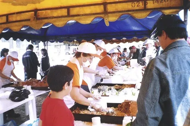 Personas mirando comida en un puesto de mercado al aire libre bajo un toldo azul