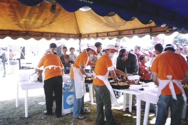 Personas con delantales naranjas sirviendo comida en mesas de bufé al aire libre bajo un toldo