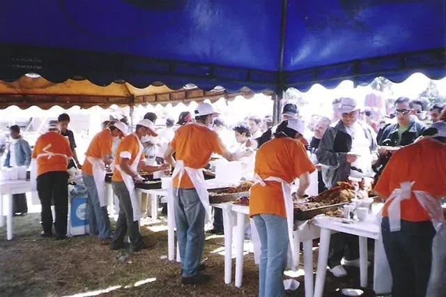 Voluntarios con delantales naranjas sirviendo comida en un evento al aire libre muy concurrido bajo una carpa azul