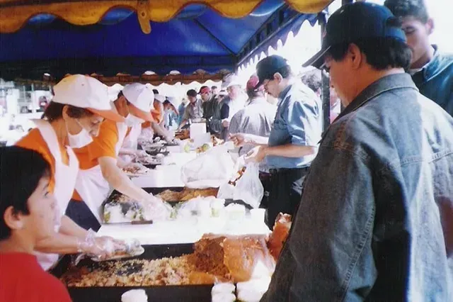 Personas sirviendo comida en una mesa de bufé bajo un toldo azul, con un hombre esperando en la fila.