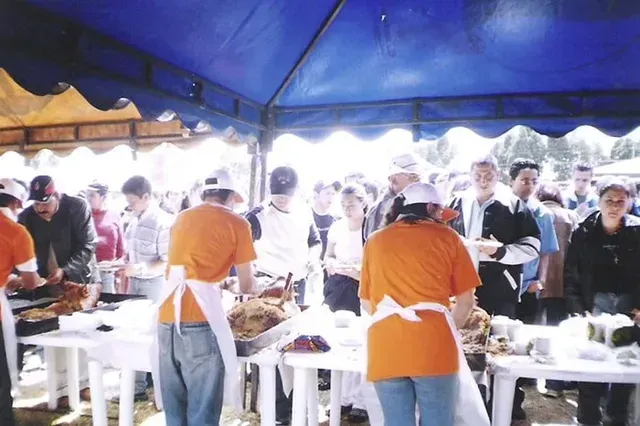 Personas sirviendo comida en un evento al aire libre muy concurrido bajo una carpa azul